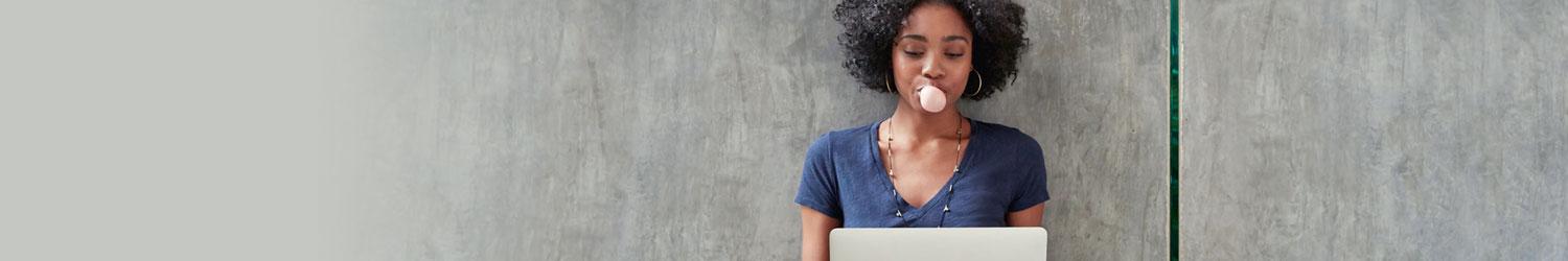 Person with curly hair blowing a bubble gum bubble while using a laptop.