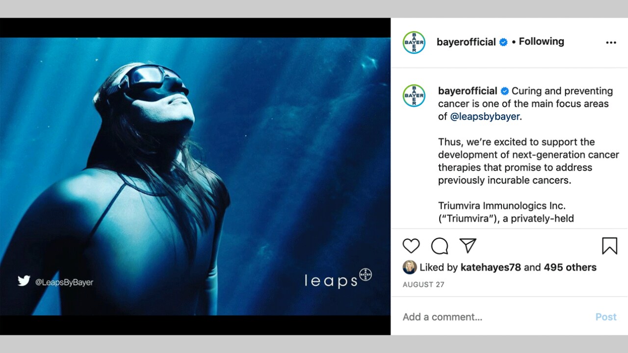 A freediver wearing goggles looks up underwater in a blue-lit scene. The caption discusses cancer research focus by Bayer, mentioning Triumvira Immunologics Inc.