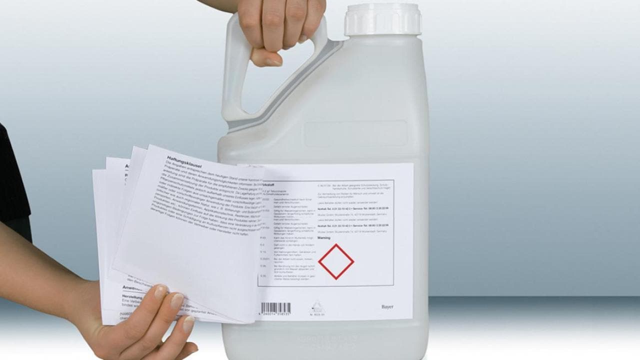 Person holding a large white chemical container with a label and a stack of safety data sheets.
