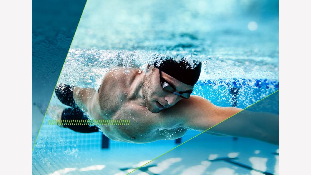 A swimmer in a black cap and goggles glides underwater in a pool, creating bubbles as he moves.