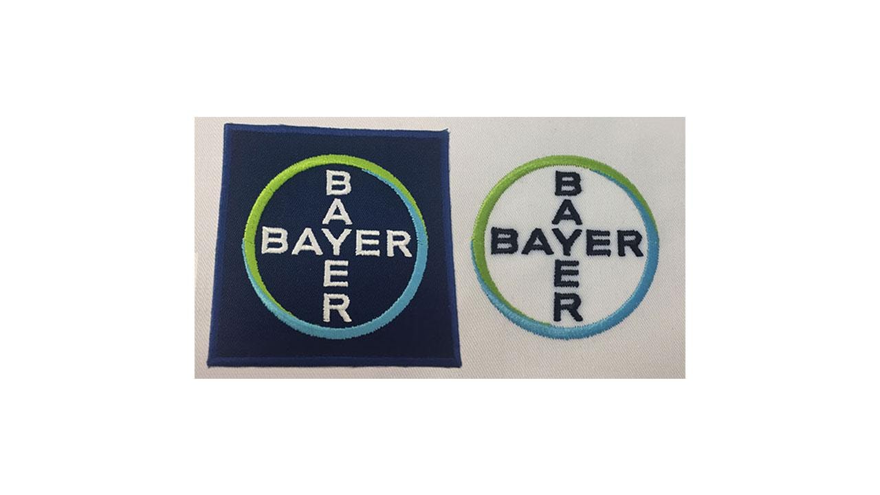 Two Bayer logo patches: one with a navy background and a circular patch with a white background. Both feature the word "BAYER" diagonally within colored rings.