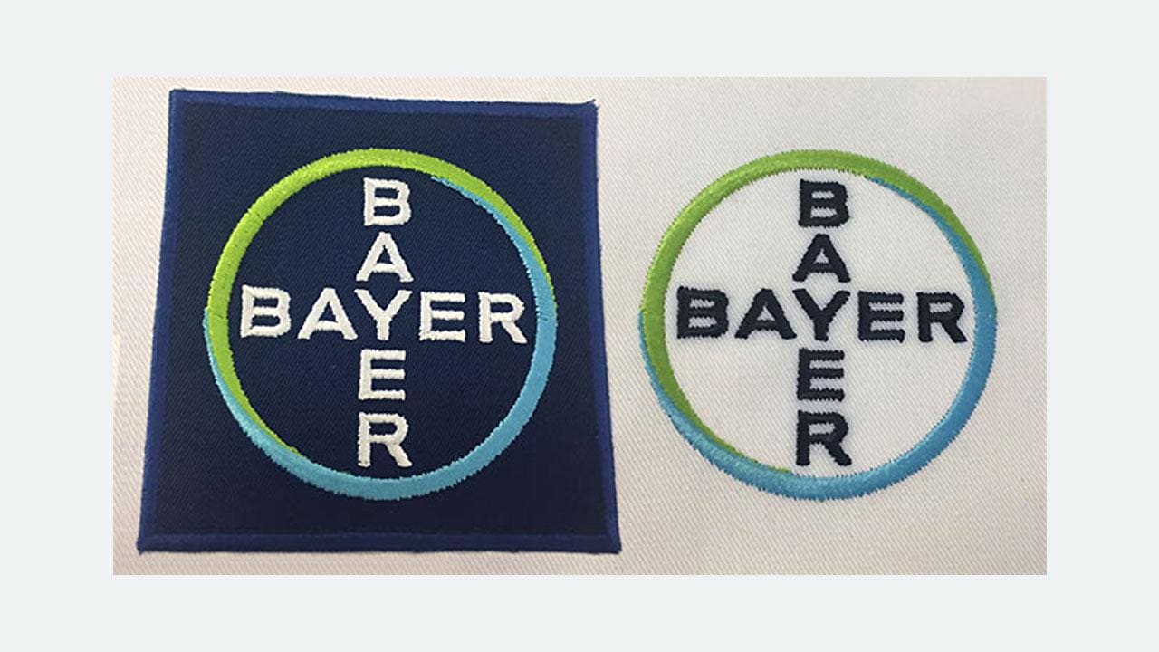 Two embroidered patches with the Bayer logo, featuring blue and green circular designs with the word "BAYER" in white. Left on a dark background, right on a light background.