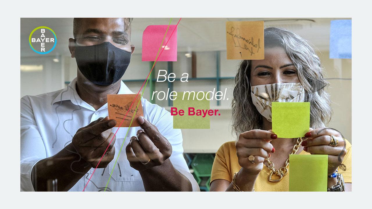 Two people wearing masks place sticky notes on glass. Text reads, "Be a role model. Be Bayer." Bayer logo is visible.