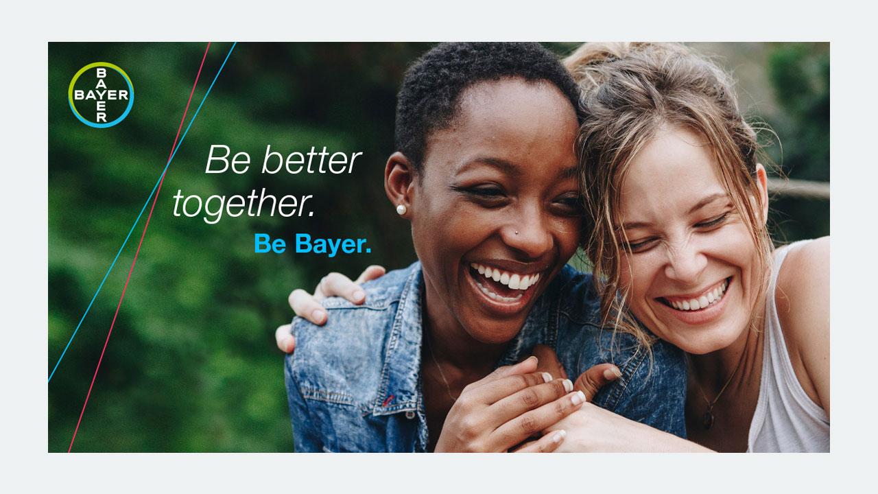 Two women smiling and embracing closely, with the text "Be better together. Be Bayer." and the Bayer logo in the top left corner.