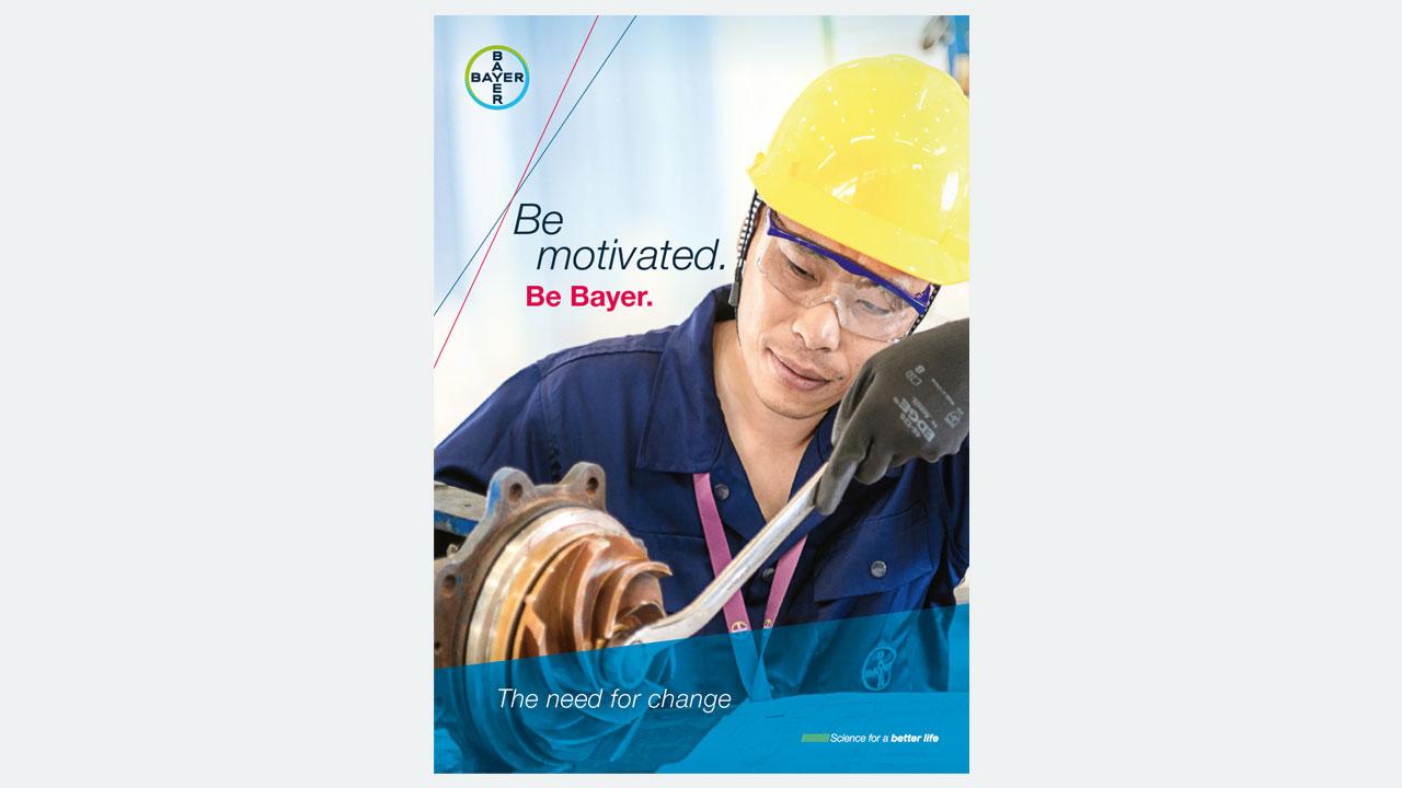 Person wearing a yellow hard hat and blue uniform examines industrial equipment. Text reads "Be motivated. Be Bayer. The need for change.