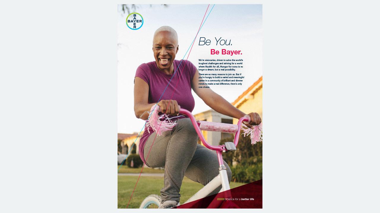 A person smiling while sitting on a bicycle with tassels, wearing a purple top and gray pants. The Bayer logo and text read: "Be You. Be Bayer.
