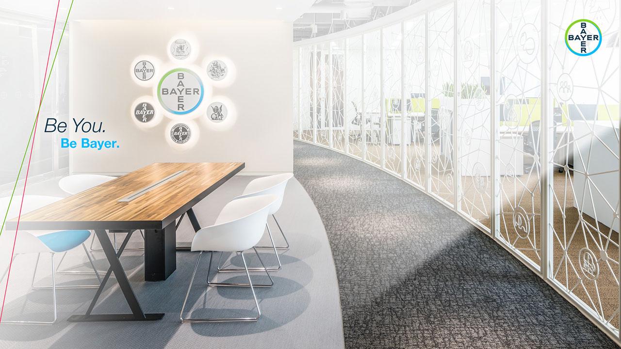 Modern office space with a long wooden table, white chairs, and decorative Bayer plates on the wall. The room features a curved wall with geometric patterns. Text reads, "Be You. Be Bayer.