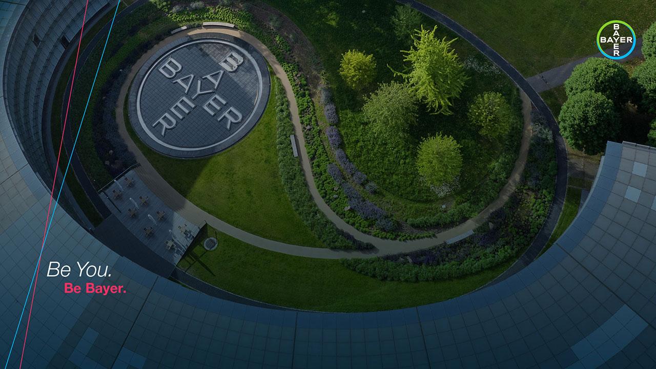 Aerial view of a corporate building courtyard with a Bayer logo on the ground. Surrounding greenery includes trees and hedges. A path curves through the landscaped area.