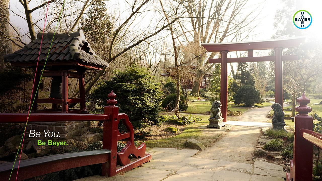A serene Japanese garden with a stone path, red wooden gate, and small statues. Trees and shrubs fill the landscape. Bayer logo in the top right corner.