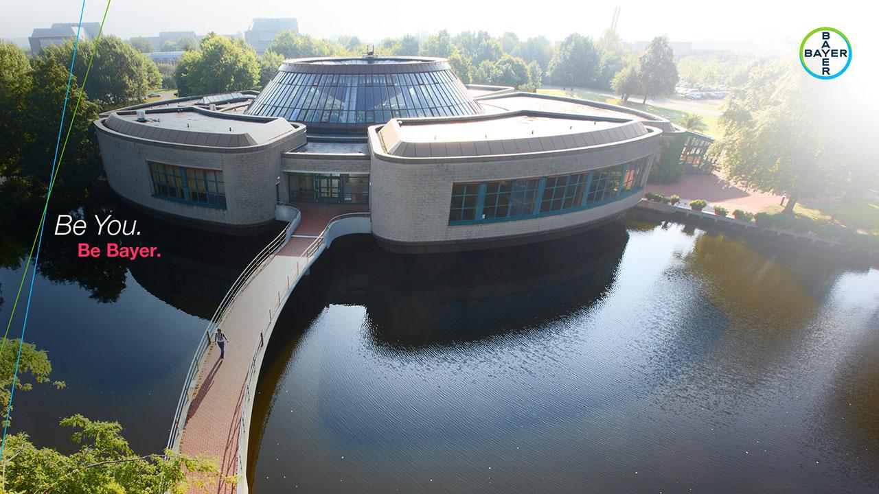 Aerial view of a circular building with a glass dome, surrounded by water and trees. A bridge leads to the entrance. The Bayer logo and slogan "Be You. Be Bayer." are visible.