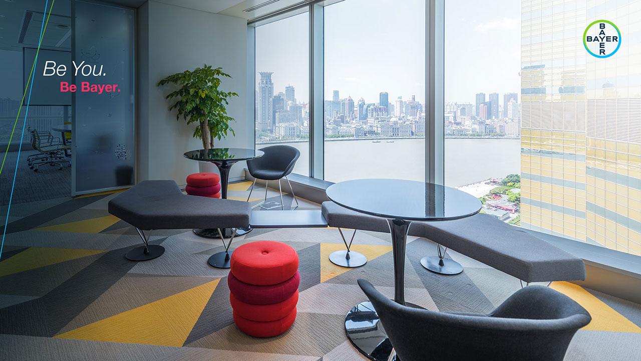 Modern office lounge with round tables, chairs, a potted plant, and a cityscape view through large windows. "Be You. Be Bayer." message on the wall.