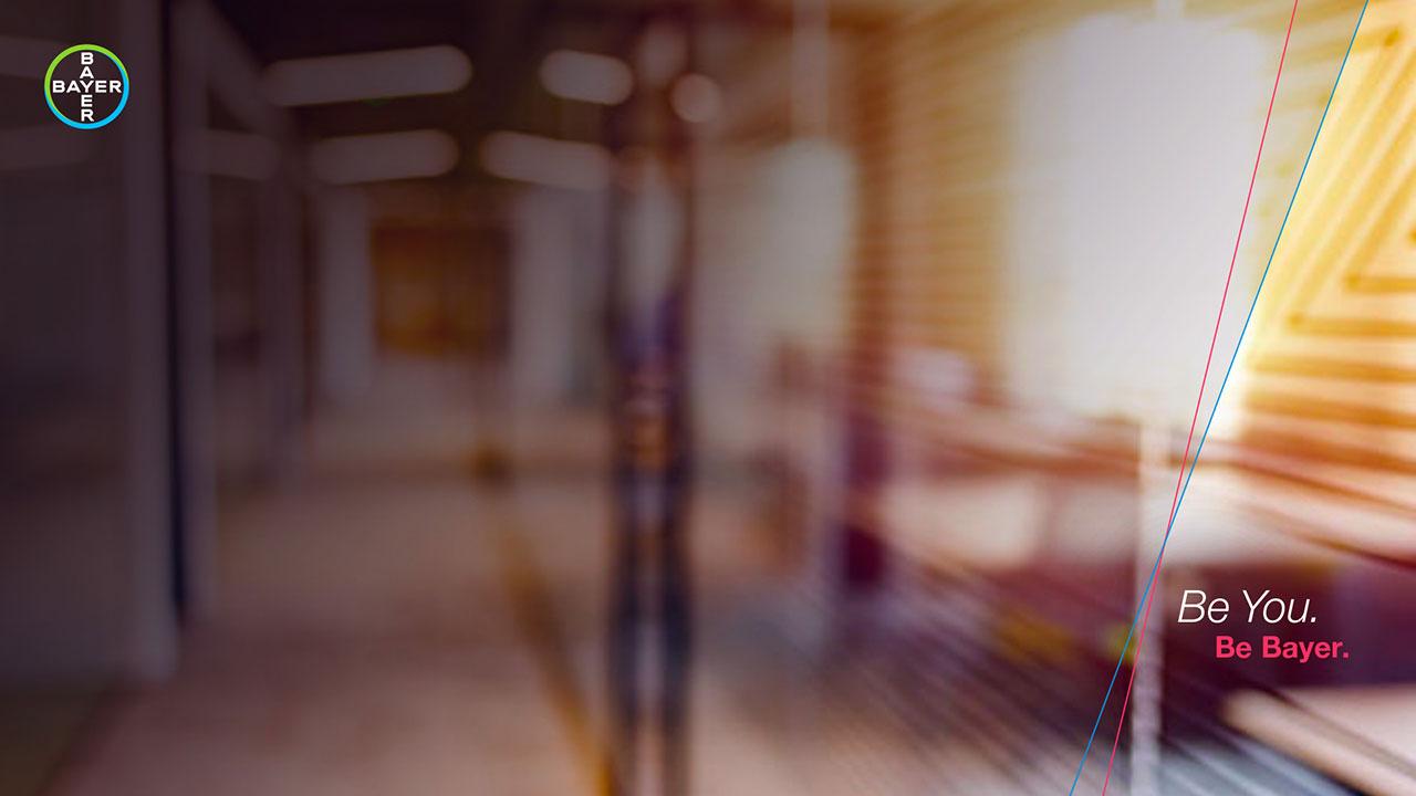 Blurred image of an office hallway with overlapping texts "Be You. Be Bayer." and a Bayer logo in the top left corner.
