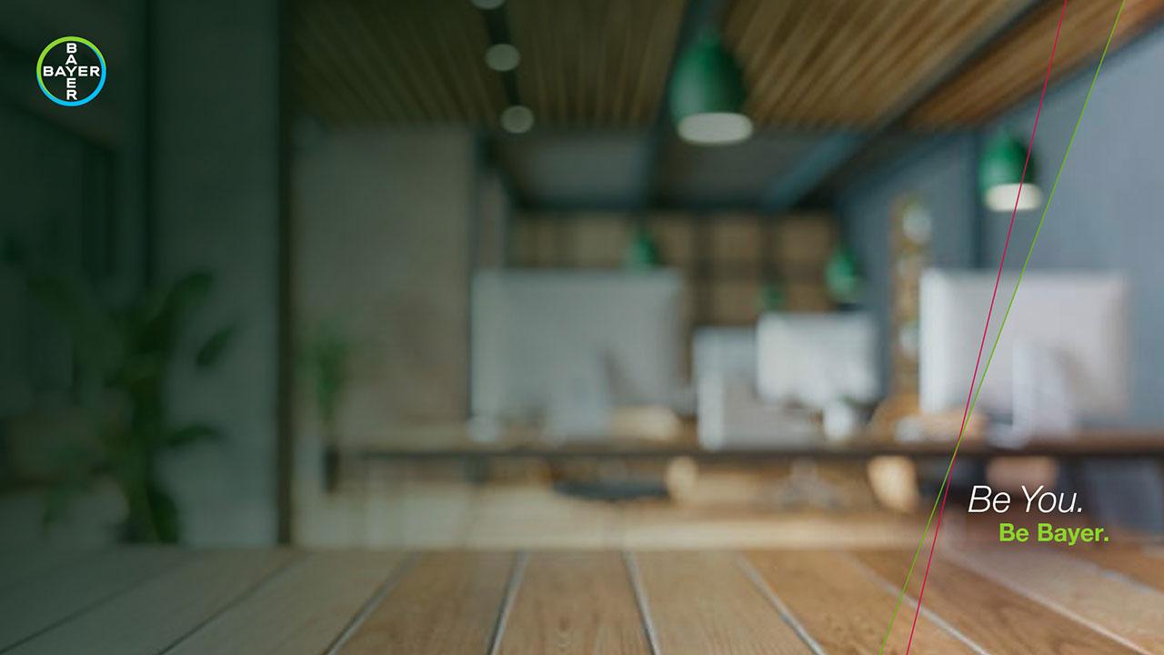Blurred office space with wooden desks and green pendant lights. The Bayer logo is in the top left corner. Text on the right reads, "Be You. Be Bayer.