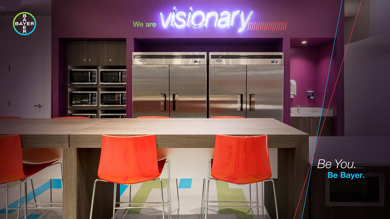 Office kitchen with purple walls, large stainless steel fridges, and a wooden table with orange chairs. Neon sign reads "We are visionary." Bayer logo and "Be You. Be Bayer." text present.