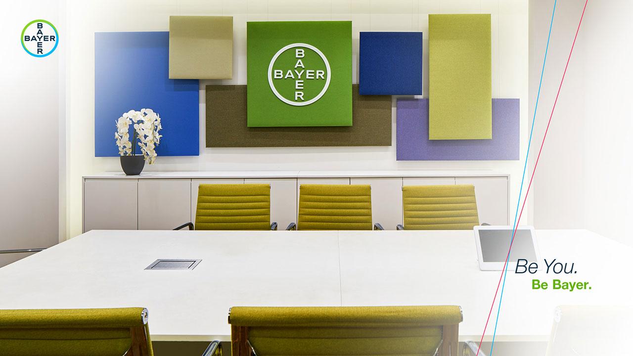 Conference room with green chairs, a table, and wall art. A prominent Bayer logo is displayed, with the slogan "Be You. Be Bayer." visible on the right side.