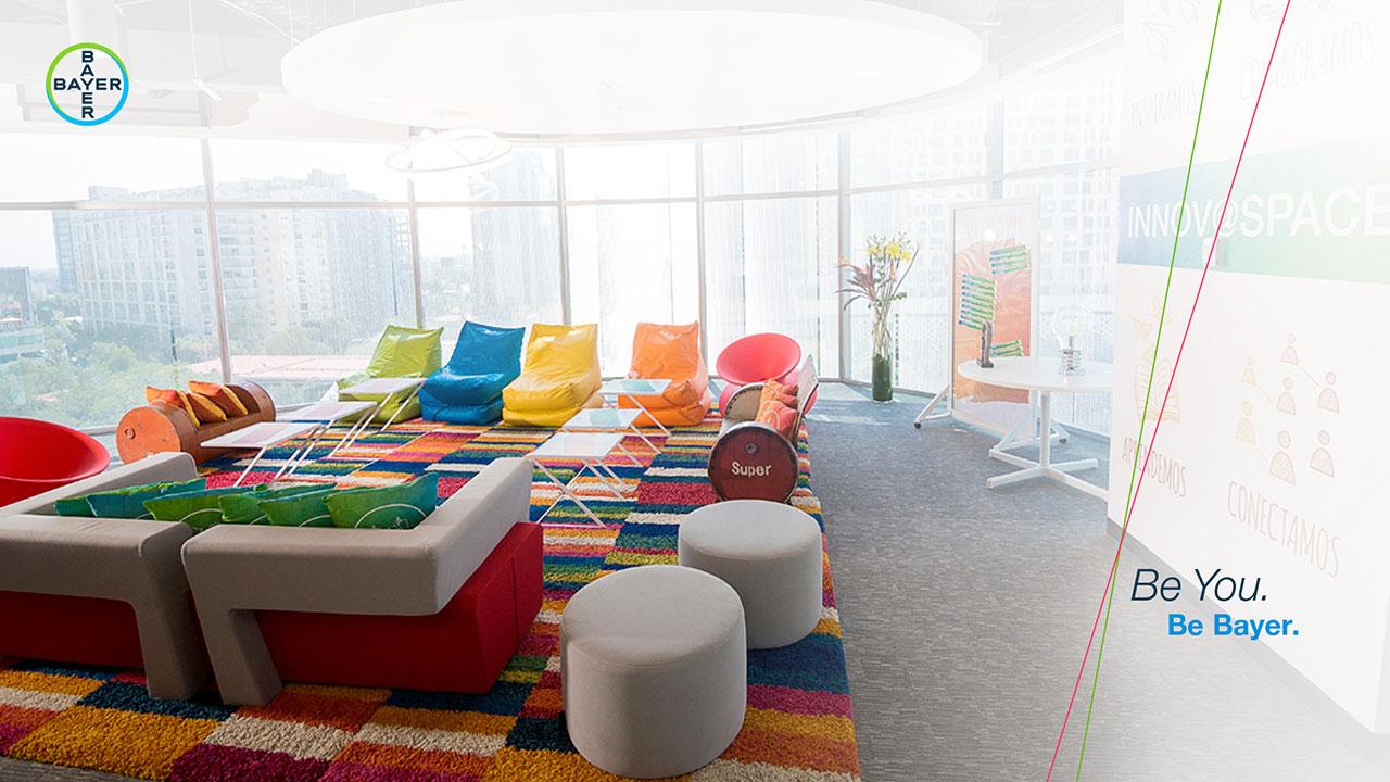 A colorful office lounge area with multicolored chairs and a vibrant rug. There are large windows and modern decor. The text reads "Be You. Be Bayer.