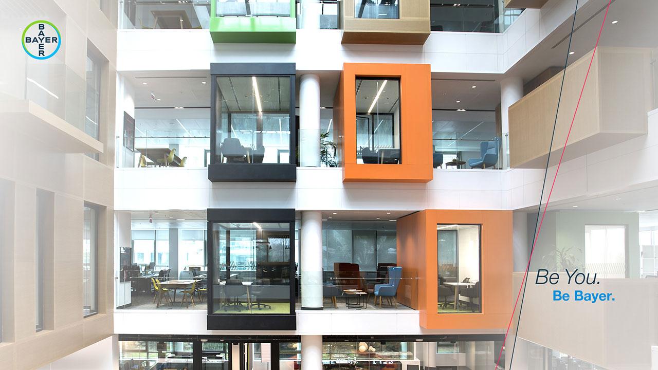 Modern office building interior with colorful, suspended meeting pods, glass walls, company logo, and text "Be You. Be Bayer.