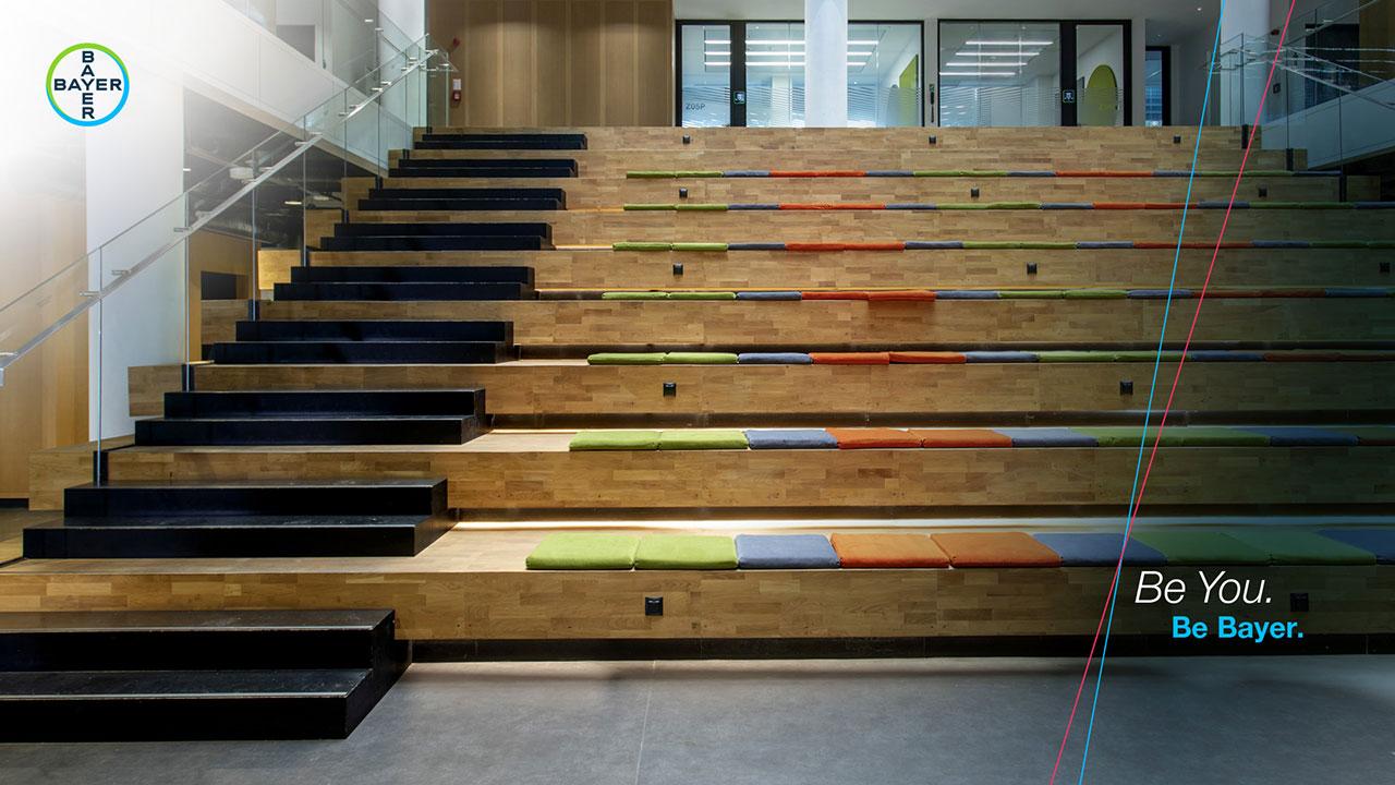 Wooden amphitheater steps with colorful cushions in an office setting. Bayer logo is in the top left corner with the text "Be You. Be Bayer." on the right.