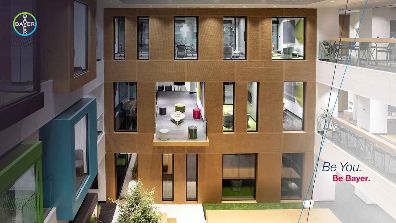 An interior view of a modern, open office space with multiple floors, glass walls, and seating areas. The Bayer logo and slogan "Be You. Be Bayer." are visible.
