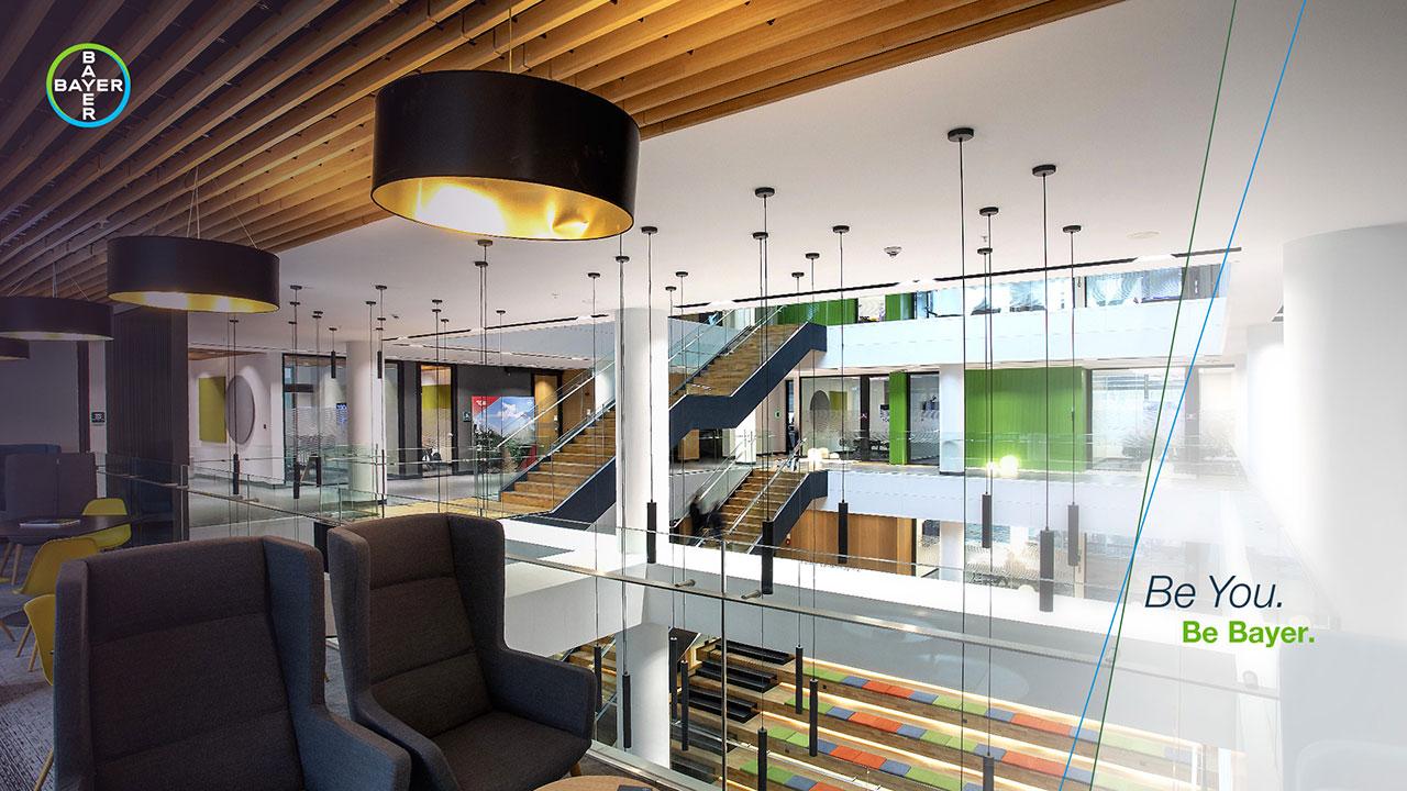 Modern office interior with glass railings, wooden ceiling accents, and multiple seating areas. Large staircase visible in background. Text reads, "Be You. Be Bayer.