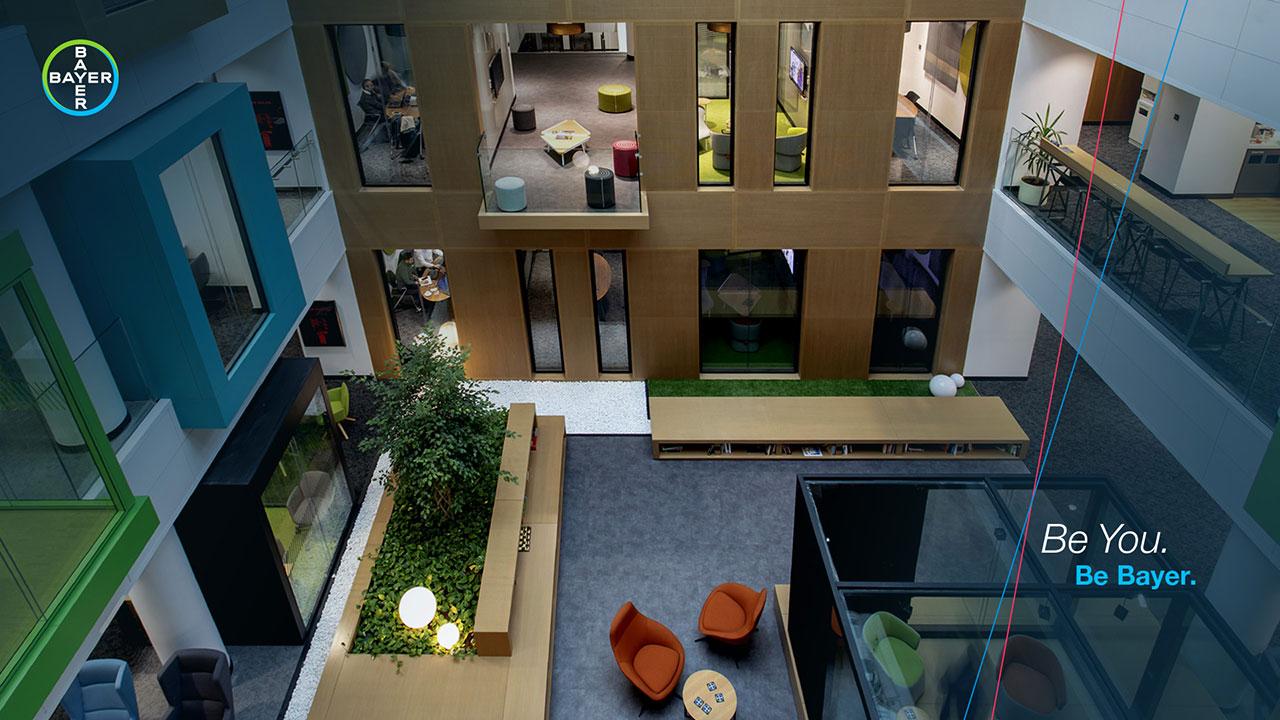 A modern office atrium with glass windows, green plants, and colorful seating areas. Bayer logo and text "Be You. Be Bayer." are visible on the side.