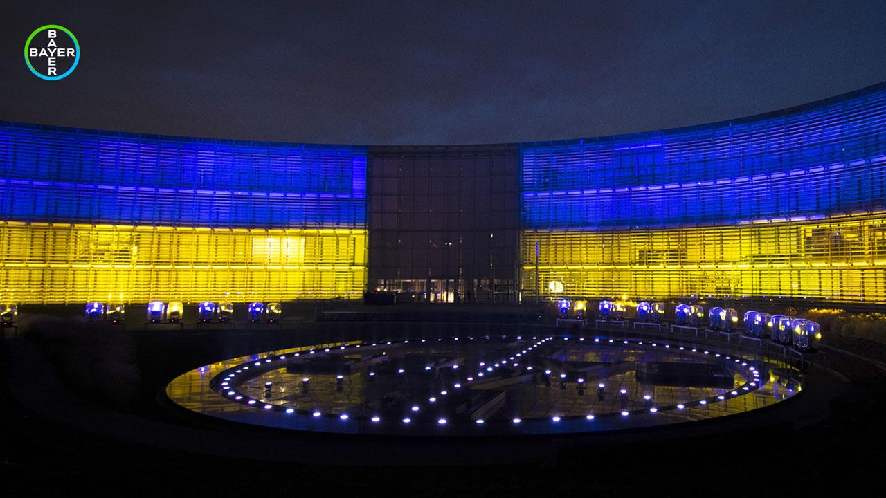 Exterior view of a large, illuminated building with blue and yellow lights. A circular area with lights is in the foreground.