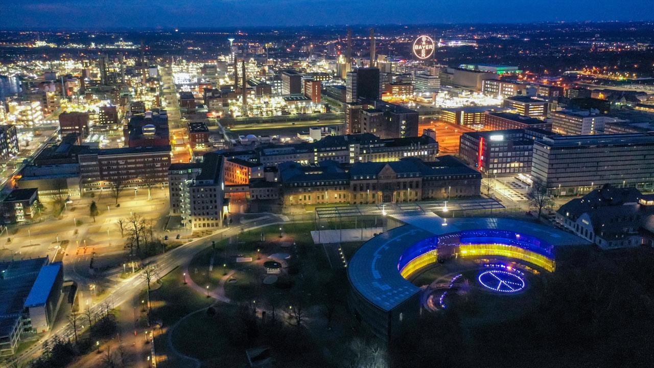 Aerial view of a cityscape at night with illuminated buildings and a circular structure displaying blue and yellow lights.