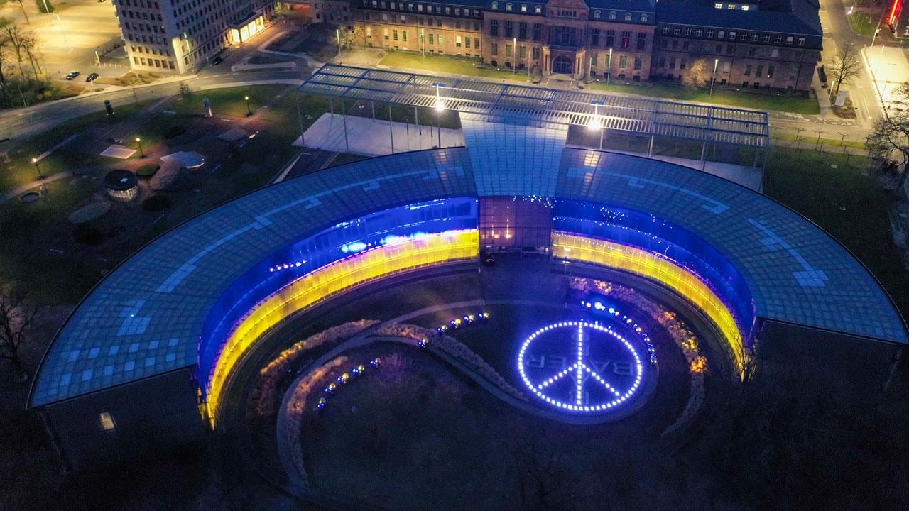 Aerial view of a horseshoe-shaped building illuminated in yellow and blue, with a large peace symbol displayed on the ground in front.