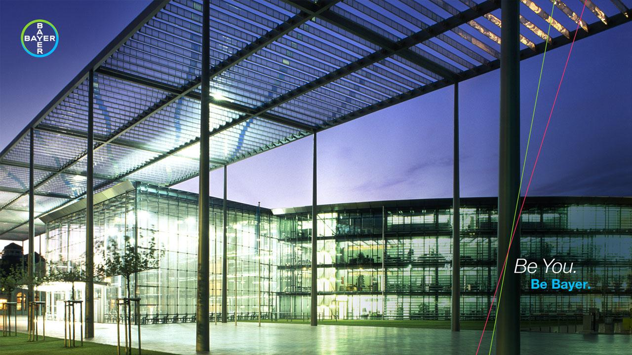Modern glass office building with illuminated interior and exterior lights at dusk, featuring the Bayer logo and slogan "Be You. Be Bayer." on the right side.