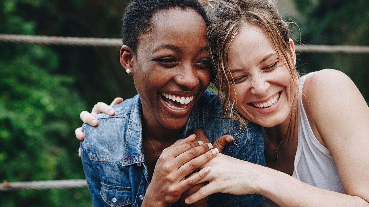 Two people laughing and embracing outdoors, with greenery in the background.