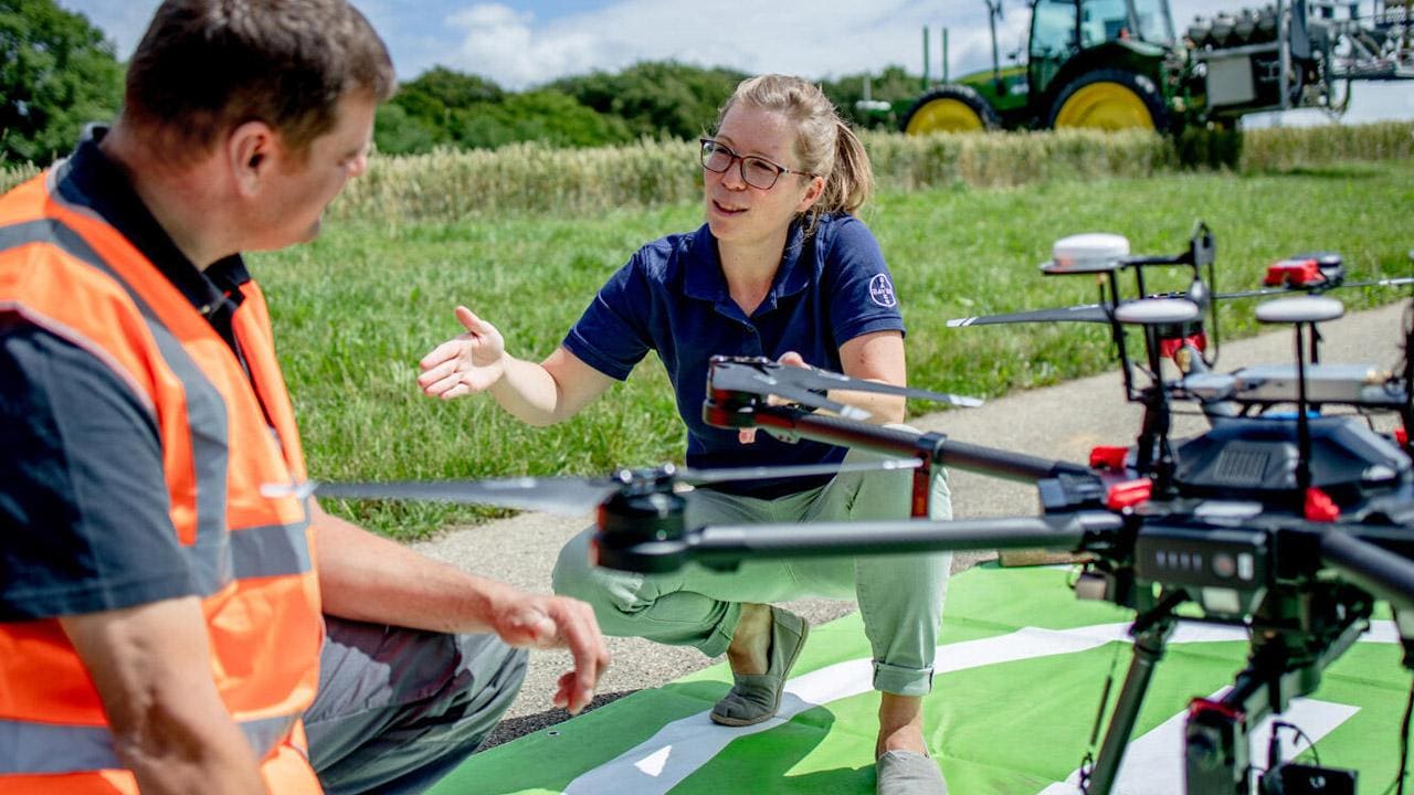 A woman and man are discussing beside a large drone on a grassy area, with a tractor visible in the background.