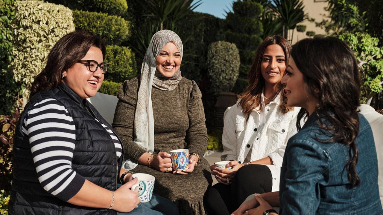 Four women sitting outdoors, smiling and holding mugs, surrounded by greenery.