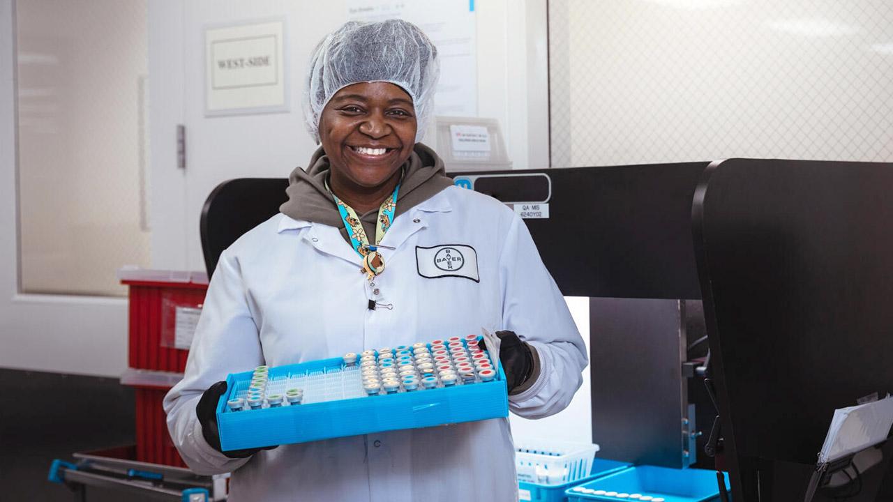 A person in lab attire smiles while holding a tray of test tubes in a laboratory setting.