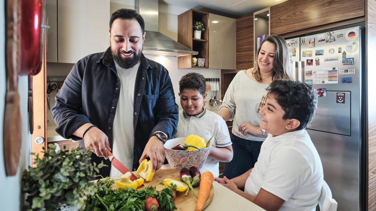 A man slices vegetables in a kitchen while two children watch and a woman stands nearby, all smiling. A bowl of chopped fruit and various vegetables are on the countertop.