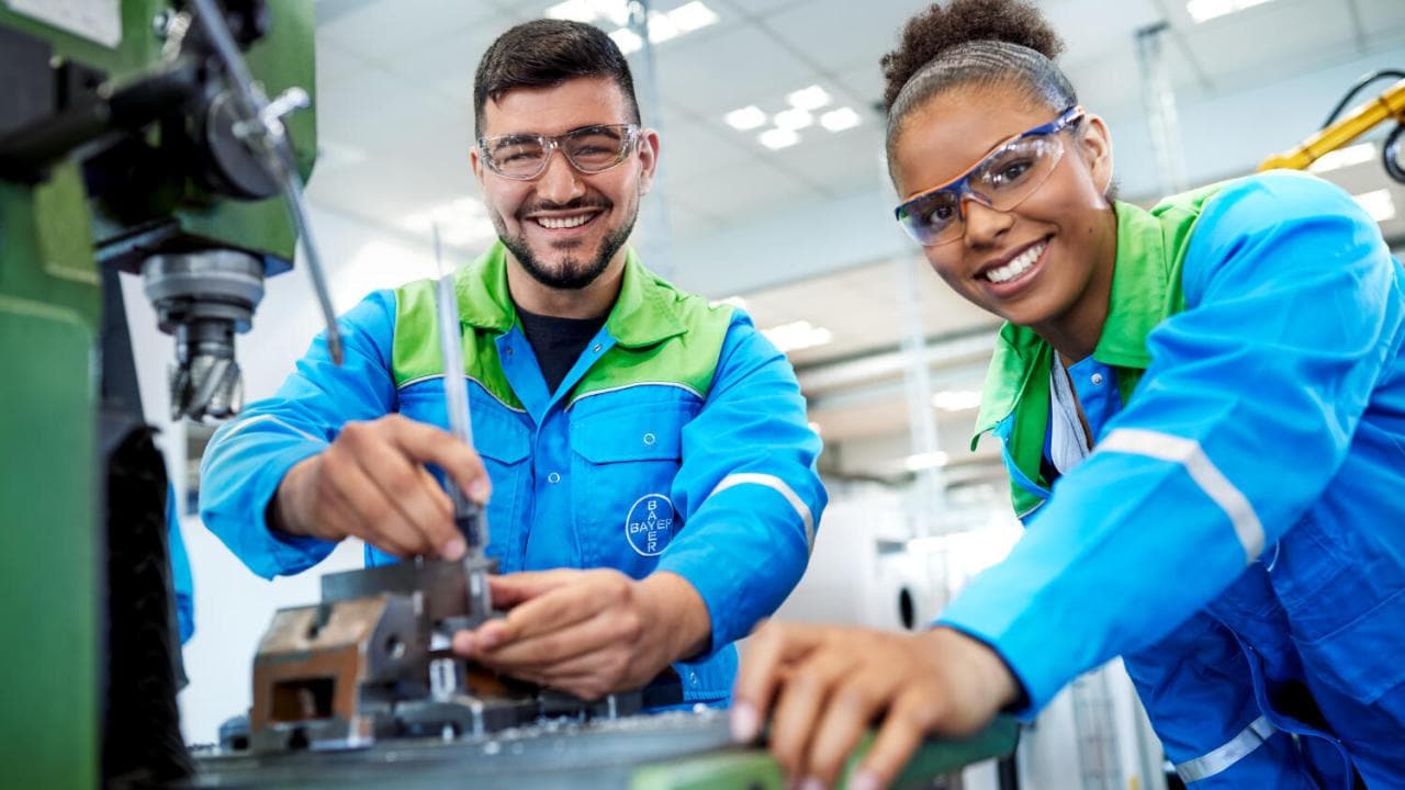 Two people in blue uniforms and safety glasses operate machinery in an industrial setting, smiling at the camera.