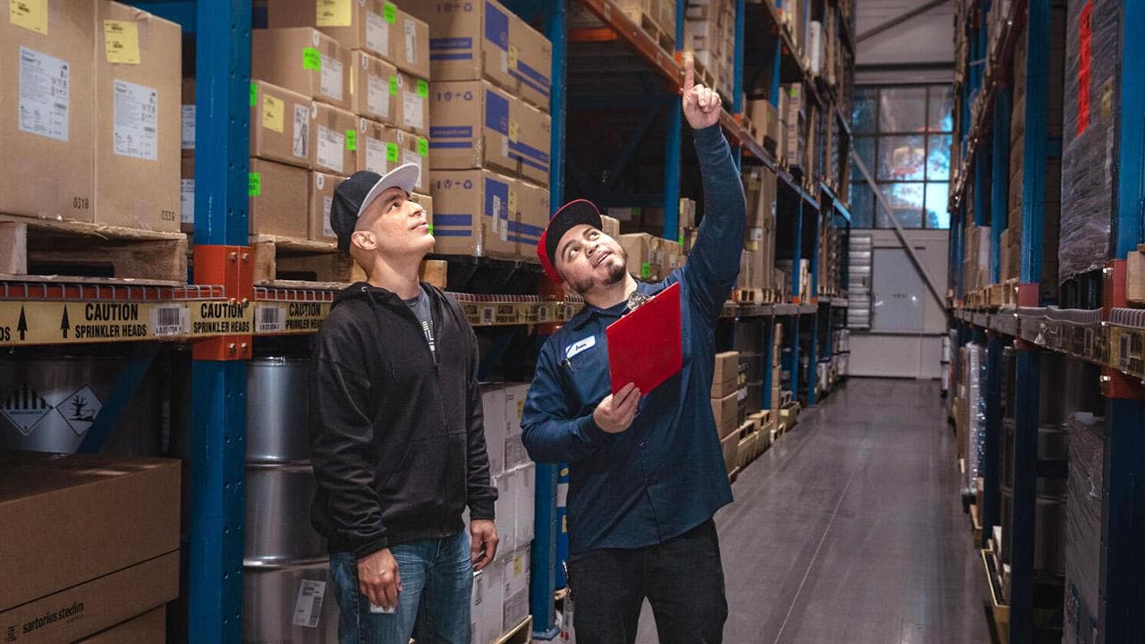 Two men wearing caps inspect boxes on tall shelves in a warehouse. One holds a clipboard and points upwards while the other looks on.