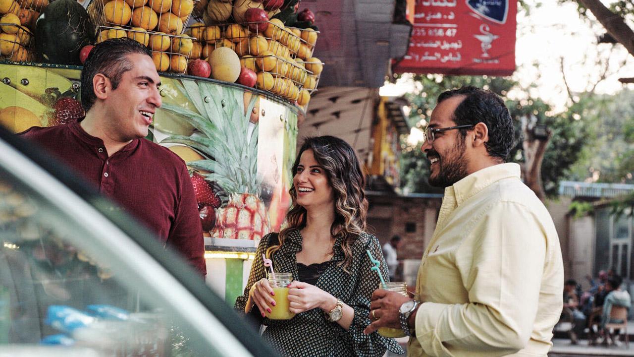 Three people stand outside a juice stand, smiling and holding drinks, with a display of colorful fruits in the background.