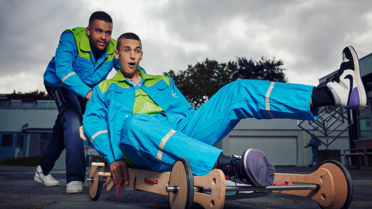 Two people in blue outfits ride a wooden cart with wheels. One is seated, excitedly steering, while the other pushes from behind, both smiling.