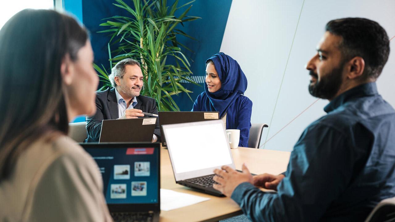 Four people in a meeting room, each with a laptop, engaged in discussion. A plant is visible in the background.