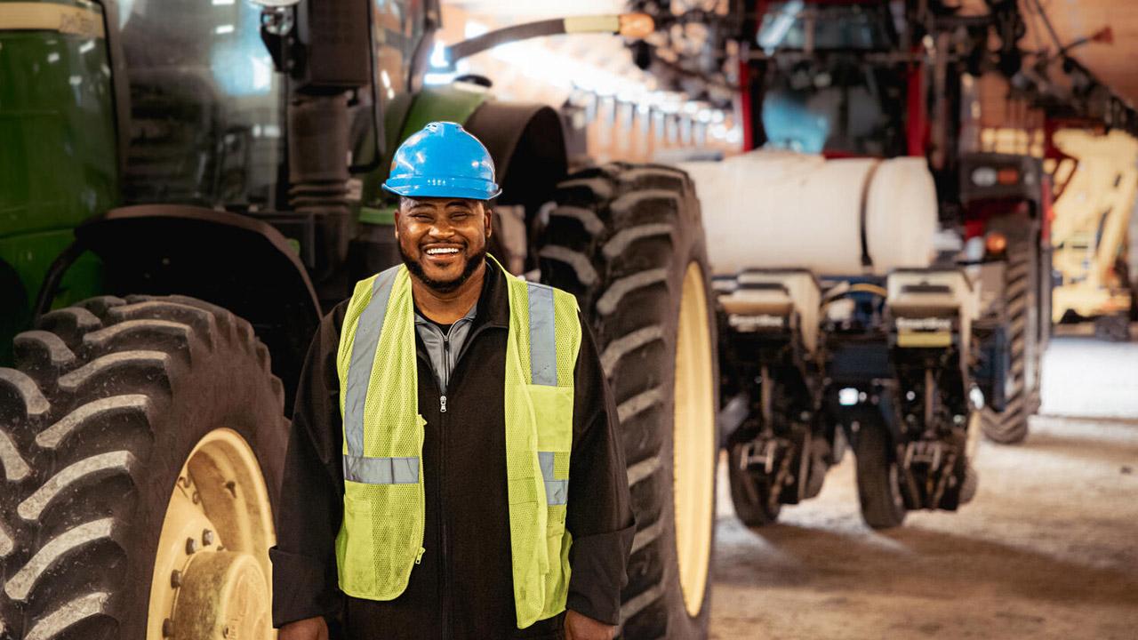 A person wearing a blue hard hat and a green safety vest stands next to large agricultural machinery indoors.