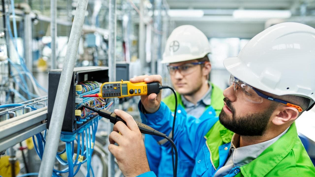 Two workers in hard hats and safety glasses use tools to inspect wiring in an industrial setting.