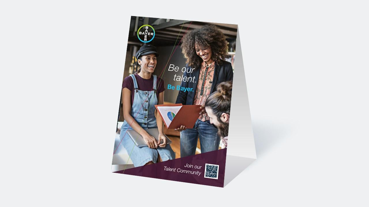 Promotional stand displaying three people smiling and looking at a laptop. Text reads, "Be our talent. Be Bayer. Join our Talent Community." Includes Bayer logo and QR code.