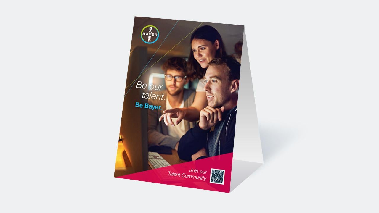 Three people working together at a computer with a Bayer recruitment poster. Text reads: "Be our talent. Be Bayer." QR code is visible.