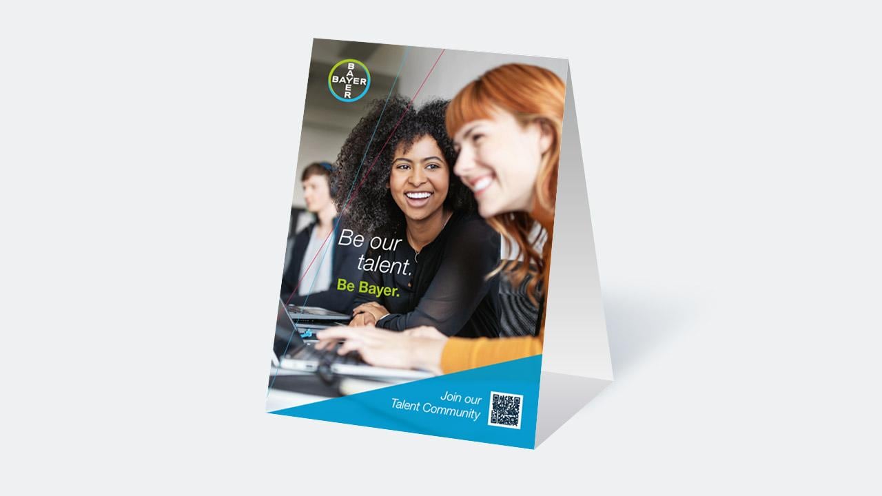 Promotional sign featuring smiling people working at laptops with text: "Be our talent. Be Bayer." and a QR code for joining the Talent Community.