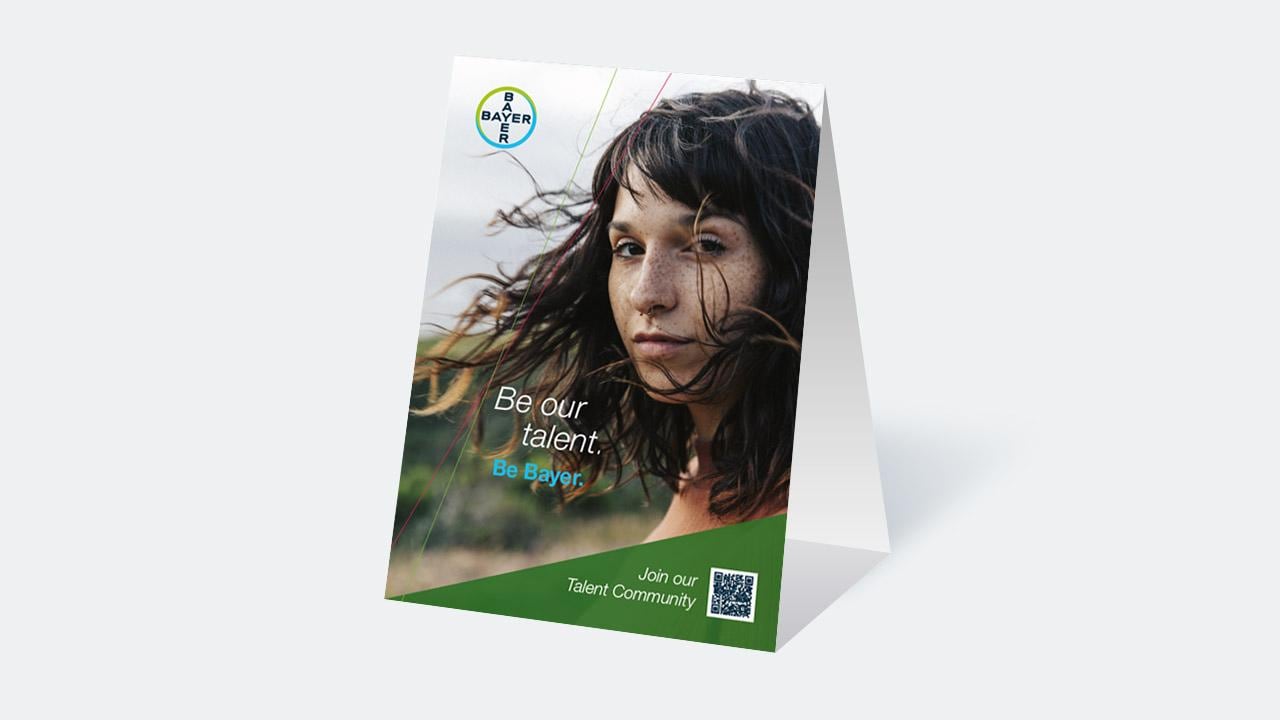 Promotional standee featuring a person with long hair, and the text "Be our talent. Be Bayer. Join our Talent Community" along with a Bayer logo and a QR code.