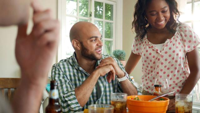A group of people sits around a table with drinks and a bowl of food, smiling and talking in a bright room.