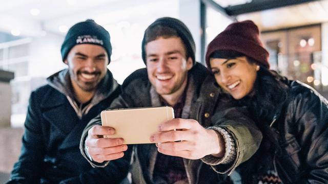Three people sitting closely together, smiling and taking a selfie with a smartphone. They are dressed warmly in jackets and beanies.