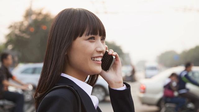 A woman in a suit smiles while talking on a cell phone outdoors, with a busy street and vehicles in the background.