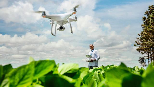 A person operates a drone over a green field, with a partly cloudy sky in the background.