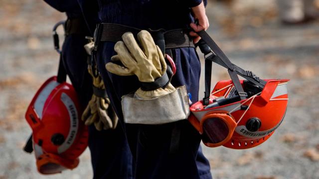 Two firefighters in gear, holding red helmets and wearing utility belts with gloves attached, standing on a gravel path with fallen leaves.